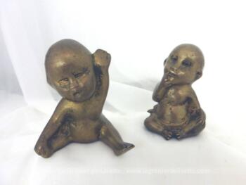 Duo de figurines de bébés en laiton de formes différentes dont le premier est allongé sur le ventre en train de sourire et le deuxième assis avec ses yeux fermés et ronds et un doigt dans la bouche.