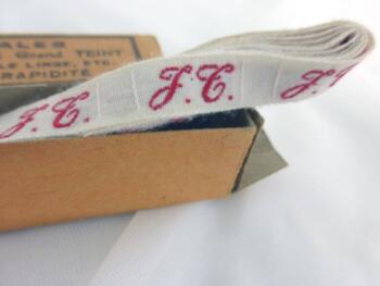 Ancien long ruban avec lettres JT brodées et sa boite en carton portant les inscriptions "Initiales Tissées Rouge Grand Teint pour marquer le linge , etc" "Economie - Rapidité".