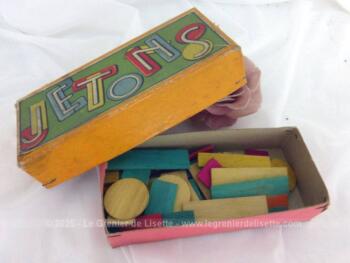 Voici une ancienne boite en carton remplie de jetons de jeu en bois de différentes formes, ronds ou rectangulaires et couleurs bleu, rouge, jaune. et vert.