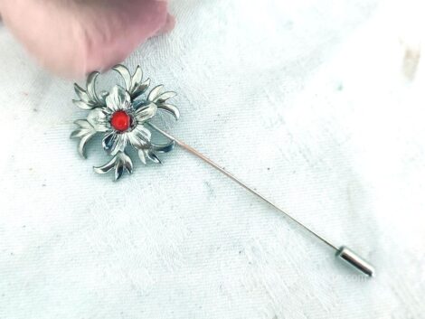 Voici une épingle à chapeaux fibule avec en décoration une fleur en métal avec une petite perle rouge au centre et son petit embout.