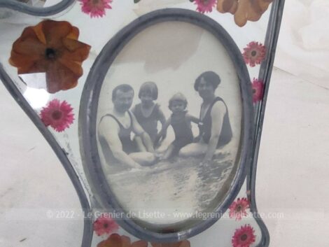 Voici un cadre en verre aux pourtours en étain façon vitrail, avec des fleurs séchées encastrées et une ancienne photo d'une famille assise au bord de l'eau.