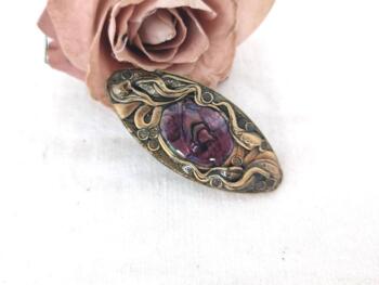 Voici une ancienne broche en laiton habillée par des décorations de guirlandes de fleurs sertissant un écusson de nacre aux tons parme.