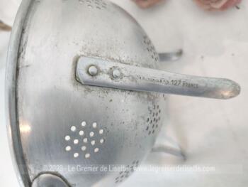 Sur 22.5 cm de diamètre, voici une ancienne passoire tripode en aluminium avec des trous pour égoutter en forme de rond et estampillée sur l'extérieur d'un pied  