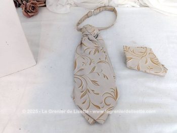 Voici une belle cravate Ascot à l'allure vintage en tissus 100% polyester beige aux dessins de volutes couleur caramel surlignées d'un liseré brillant avec une pochette assortie.