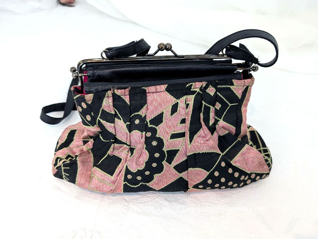 De la marque "Emma Blonde", voici un superbe sac minaudière vintage en tissu jacquard noir et rose très Art Déco, avec un haut en cuir noir et fermé par une armature en métal et doublé d'un satin fuchsia. A porterà l'épaule grâce à sa bandoulière en cuir noir ou à la main. Années 2000.